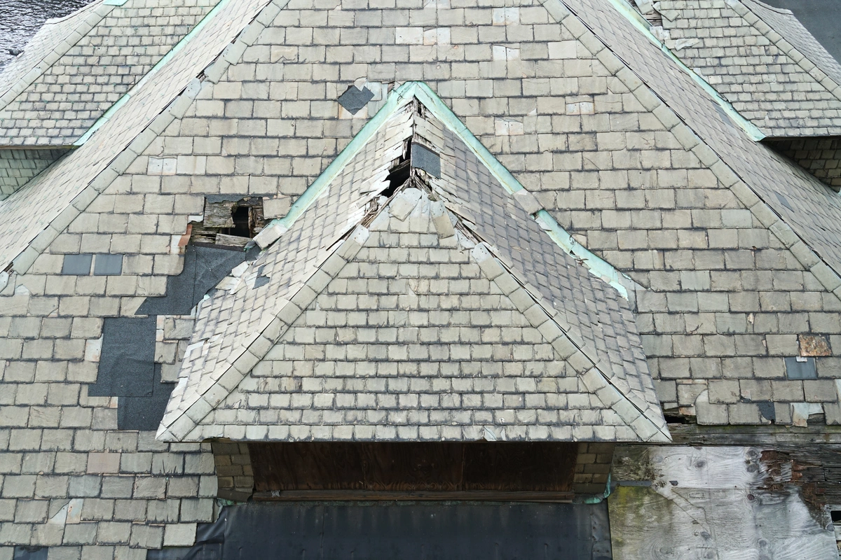 A damaged roof covered in grey shingles with visible holes, missing and broken shingles, and some areas patched with black material—signs of neglect that may leave you wondering, how old is my roof?.