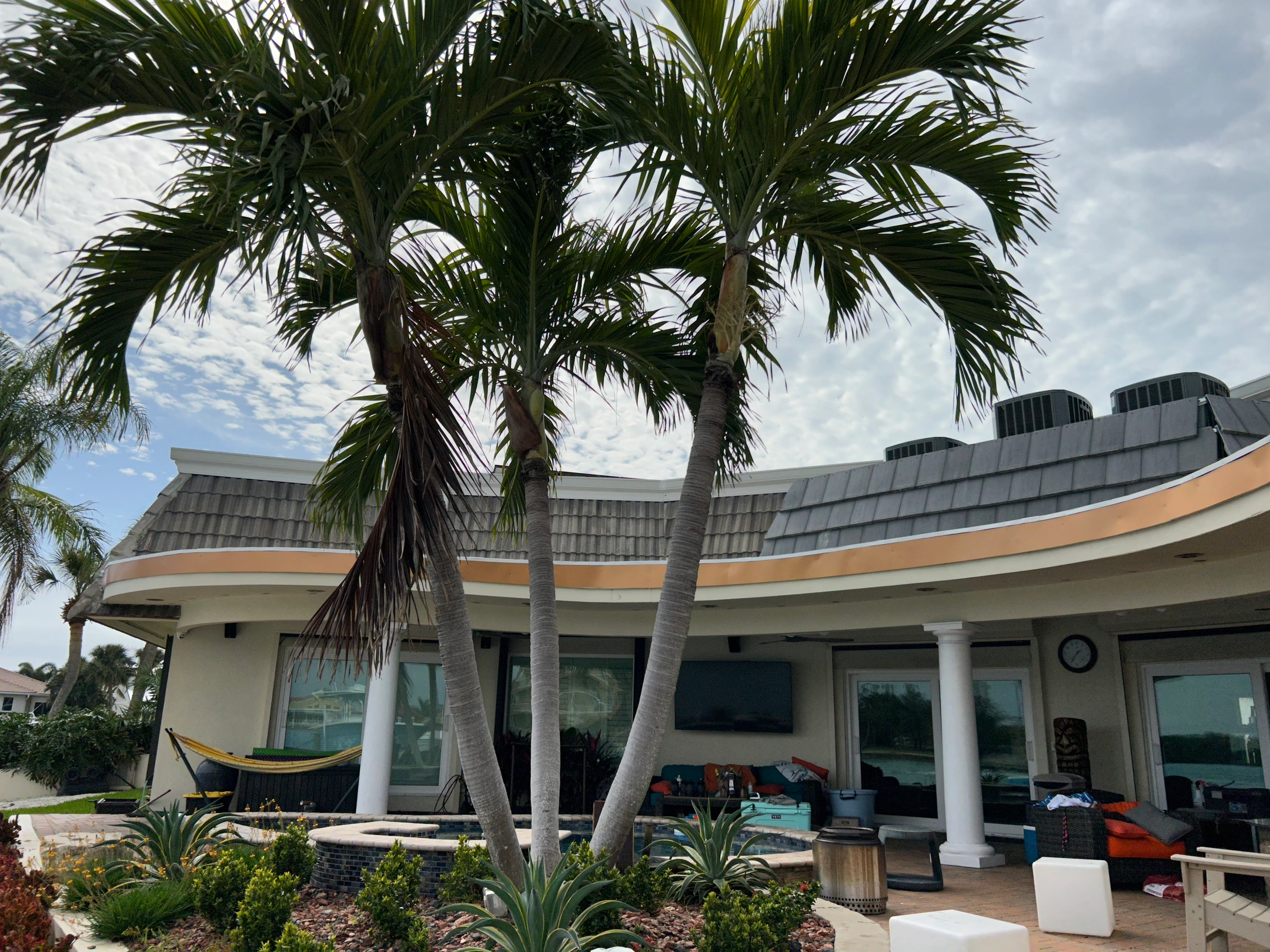 A modern house with large windows and a shaded patio area is seen behind two tall palm trees and landscaped greenery under a partly cloudy sky. Patio furniture and a hammock are visible.