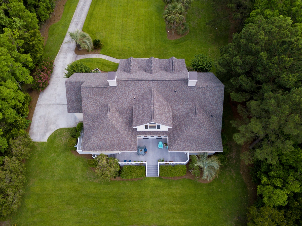 Aerial view of a large house with a gray roof featuring Tamko shingles, surrounded by green grass, trees, and bushes. A driveway curves to the left, and there’s patio furniture on the front porch—a perfect scene for a Tamko shingles review.