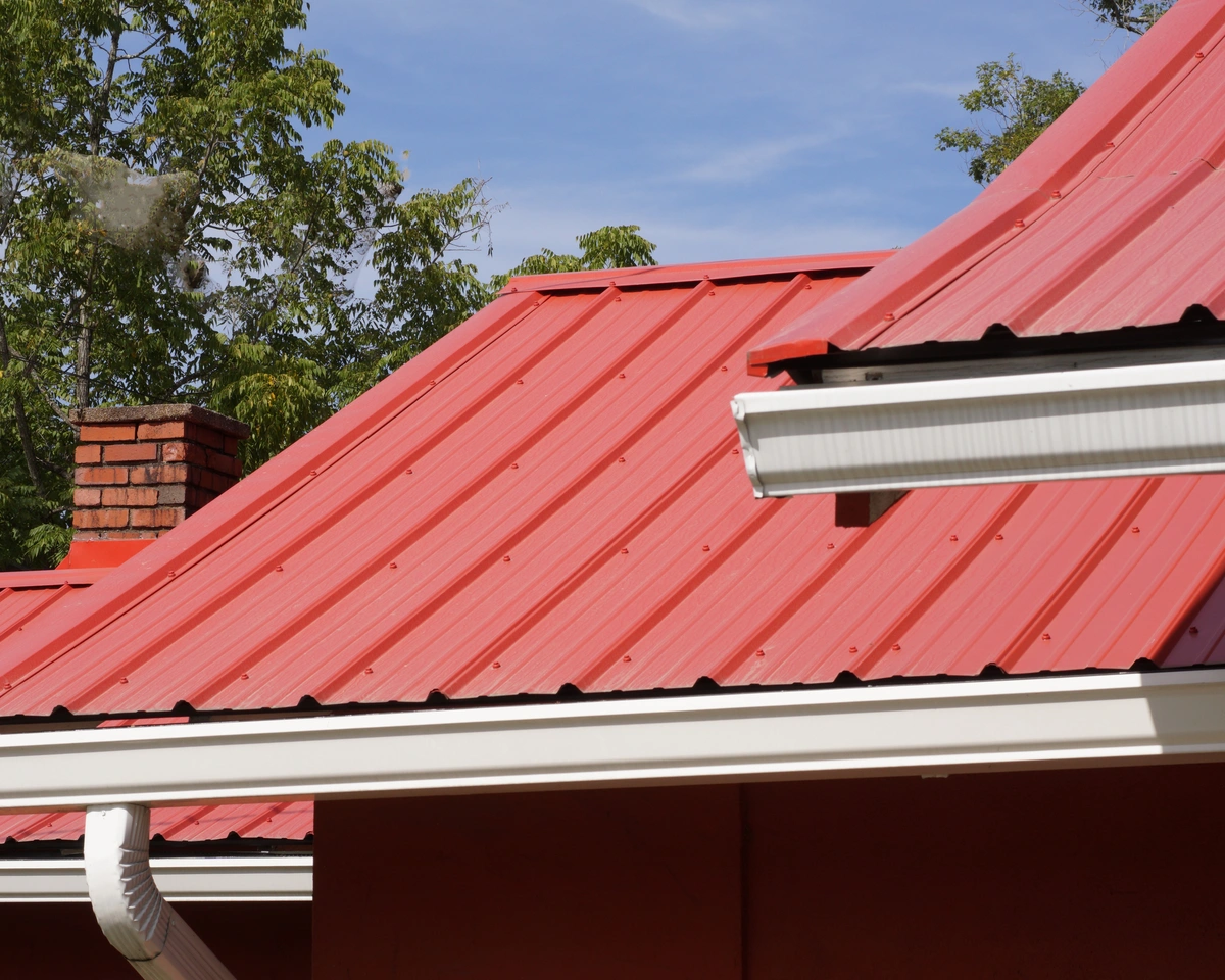 A red metal roof with visible ridges, showcasing one of the popular types of metal roofing, features white gutters on a building. A brick chimney rises amid a blue sky and green trees in the background.