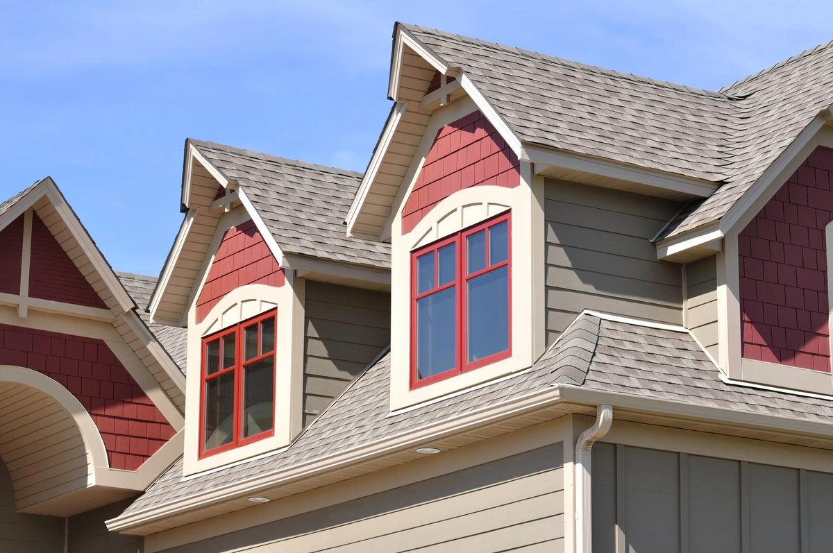 Close-up of a house exterior featuring three peaked dormer windows with red trim, beige siding, and gray architectural shingles on the roof under a clear blue sky.