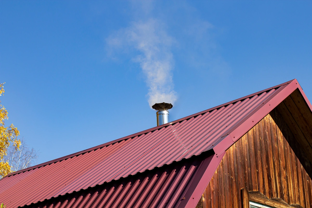 Smoke rises from a metal chimney on the standing seam metal roof of a wooden house, set against a clear blue sky. Part of a yellow-leaved tree is visible on the left edge of the image.