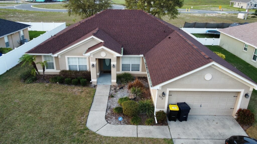 Aerial view of a single-story beige house with a brown roof, attached two-car garage, small front porch, and a curved walkway. The yard features sparse landscaping and two trash bins near the garage.