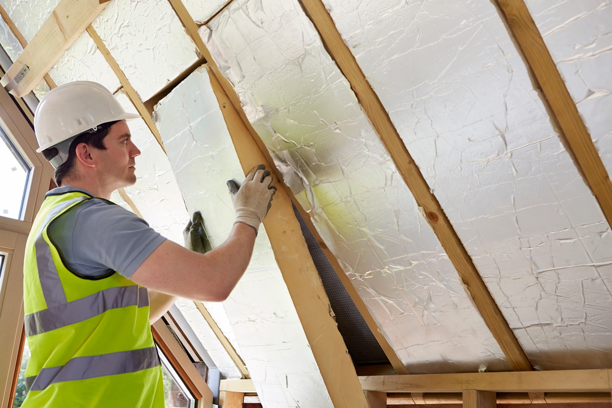 A construction worker wearing a hard hat, gloves, and a high-visibility vest installs roof insulation by fitting foil-backed panels between wooden beams in an attic.