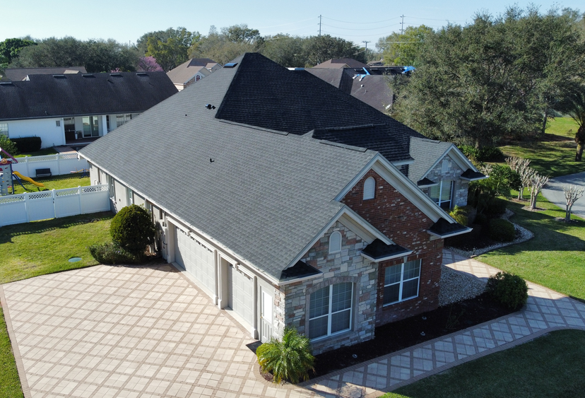 Aerial view of a single-story house with a dark gray roof, brick and stone exterior, multiple windows, and a large patterned driveway, surrounded by grass and neighboring houses.