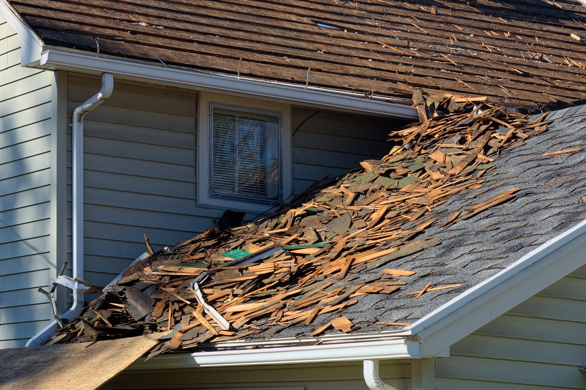 A house with piles of broken shingles and debris on the lower roof, likely from an ongoing roof tear off or replacement project; the upper section of the roof is exposed and partially stripped.