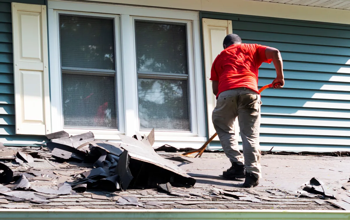 A person in a red shirt and tan pants demonstrates how to remove shingles, using a tool to lift old roofing near a window with white shutters and blue siding.