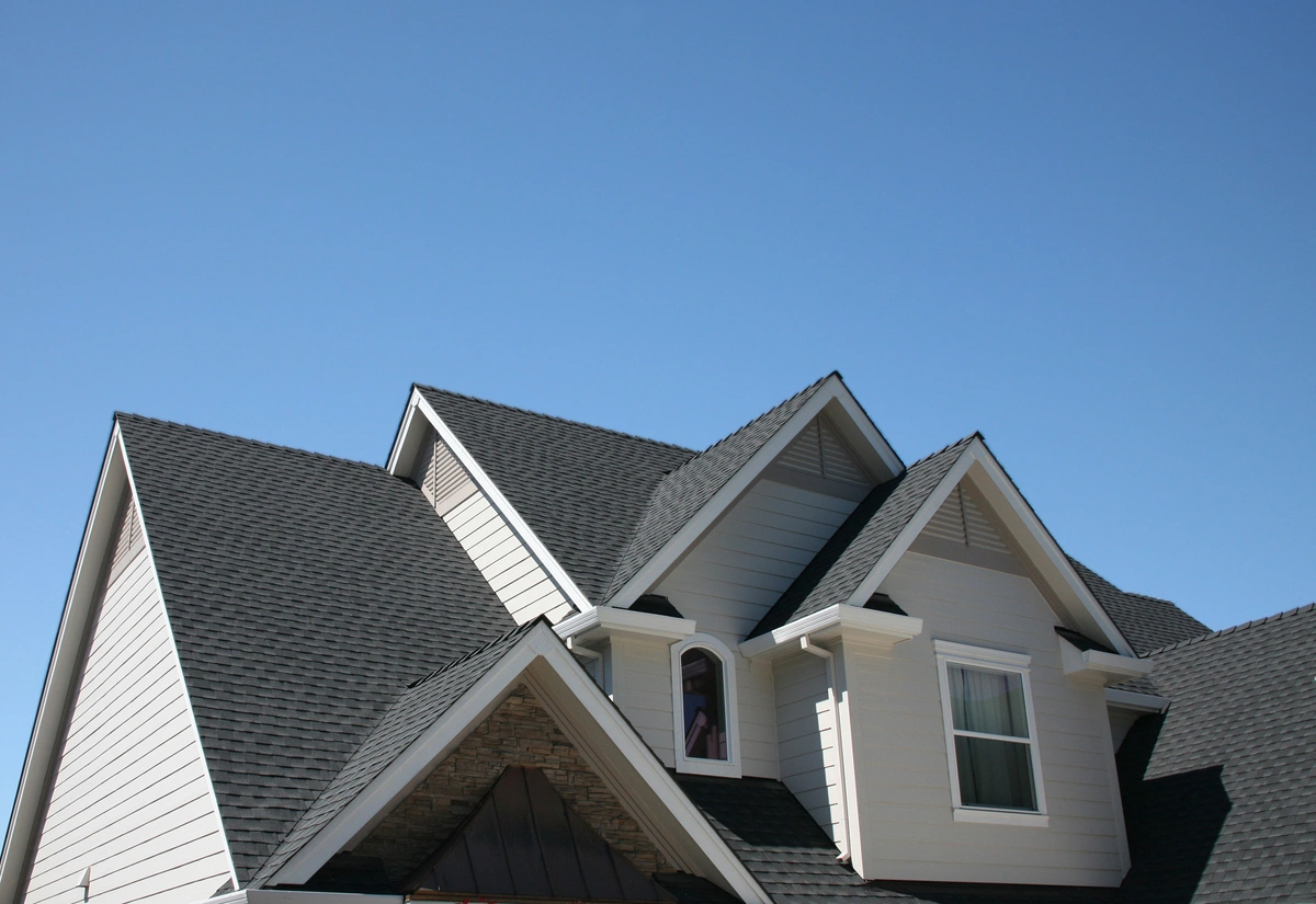 A close-up of a house roof with multiple peaks highlights the parts of a roof, featuring gray shingles, white siding, and a clear blue sky in the background.