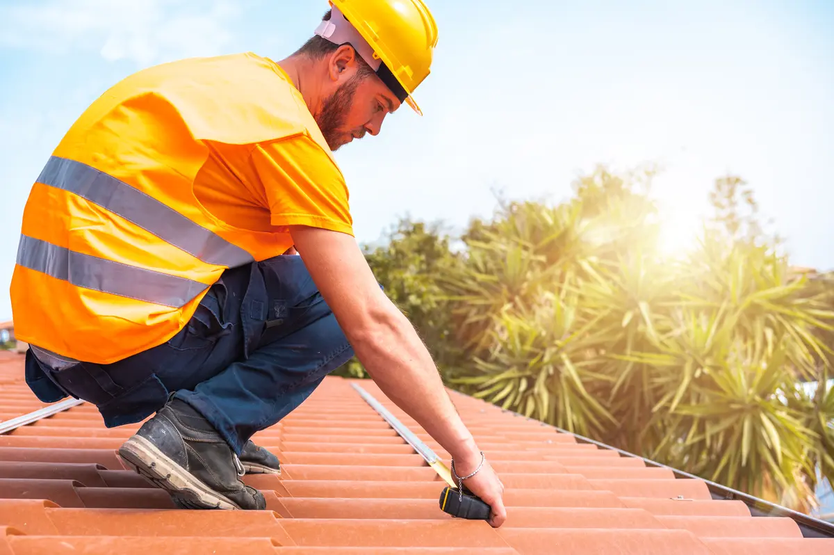 A construction worker in an orange safety vest and yellow hard hat measures a red tile roof with a tape measure, demonstrating how to calculate roof pitch on a sunny day, with trees visible in the background.