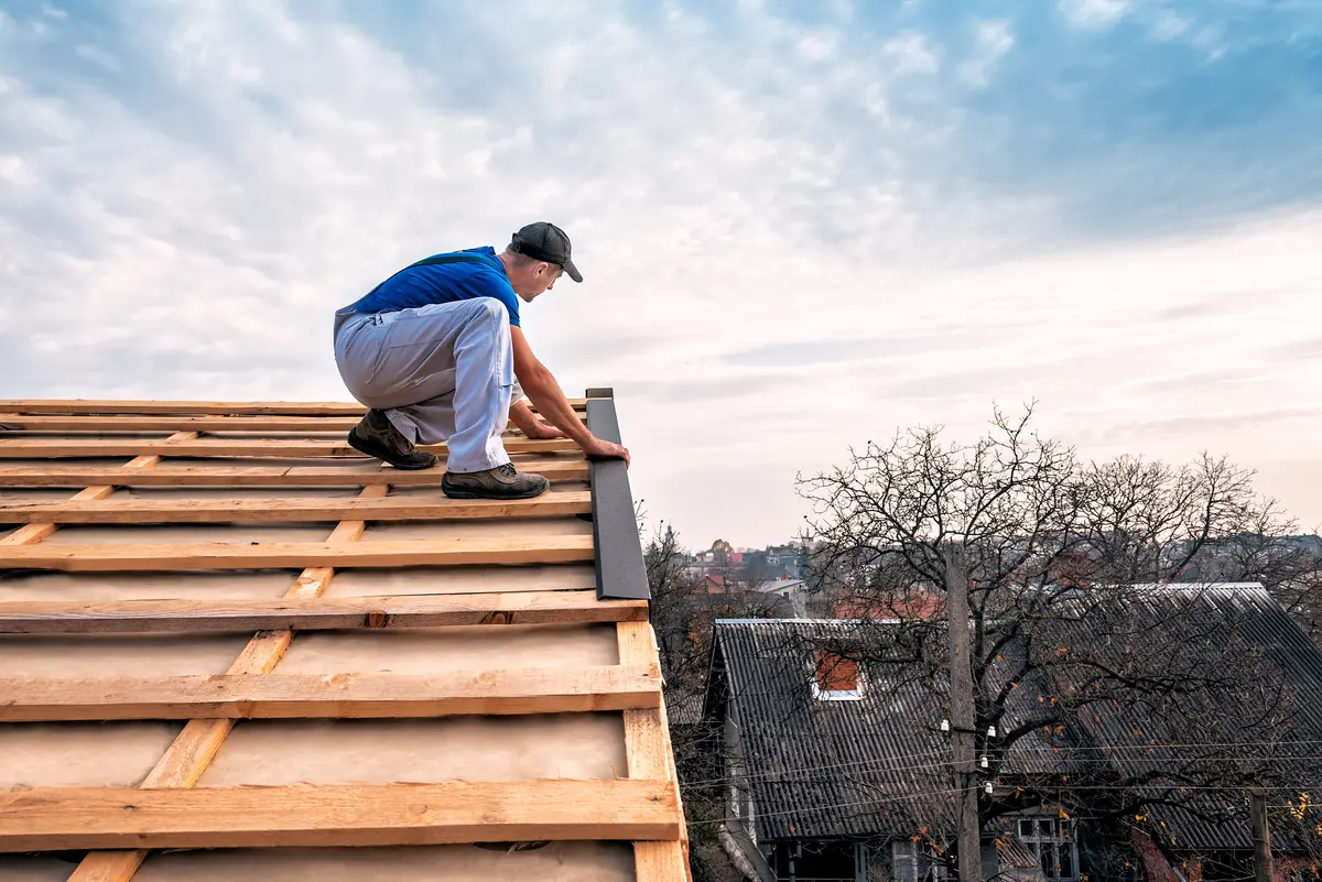A worker in a blue shirt and cap installs roof sheathing on the wooden frame of a roof, with houses and leafless trees visible in the background under a cloudy sky.
