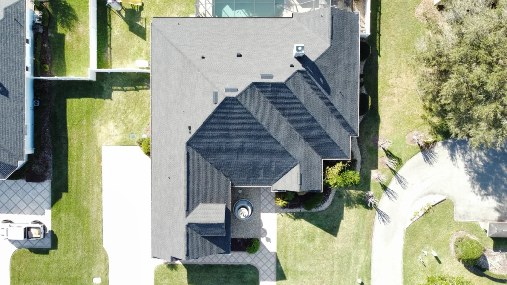 Aerial view of a large house with a dark, multi-angled roof, expertly crafted by roofing Pinellas County specialists, surrounded by green lawns, a driveway, and neighboring houses. The yard has neatly trimmed grass and paved walkways.