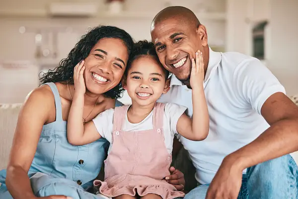 A smiling family of three, including a young girl sitting between her parents, sits close together on a couch at home in Thonotosassa. The parents and child are all smiling and holding each other's faces affectionately, feeling safe under their quality roofing.