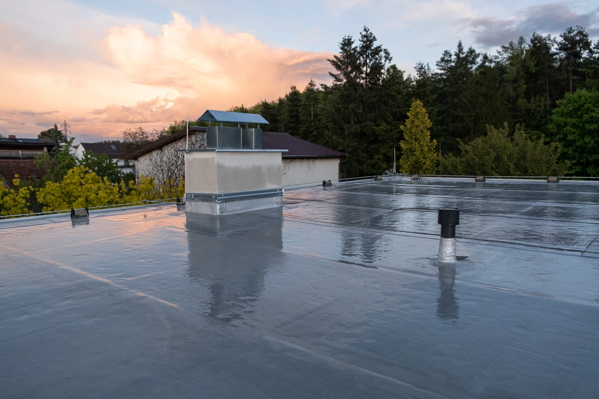 A flat, reflective rooftop featuring vents and a chimney hints at recent rubber roof repair, surrounded by trees and houses beneath a partly cloudy sunset sky.