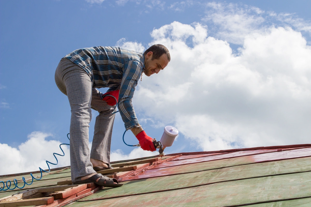 A man in a plaid shirt and gray pants uses a spray gun to apply metal roof coating, painting a roof red while standing on a sloped surface under a blue sky with clouds.