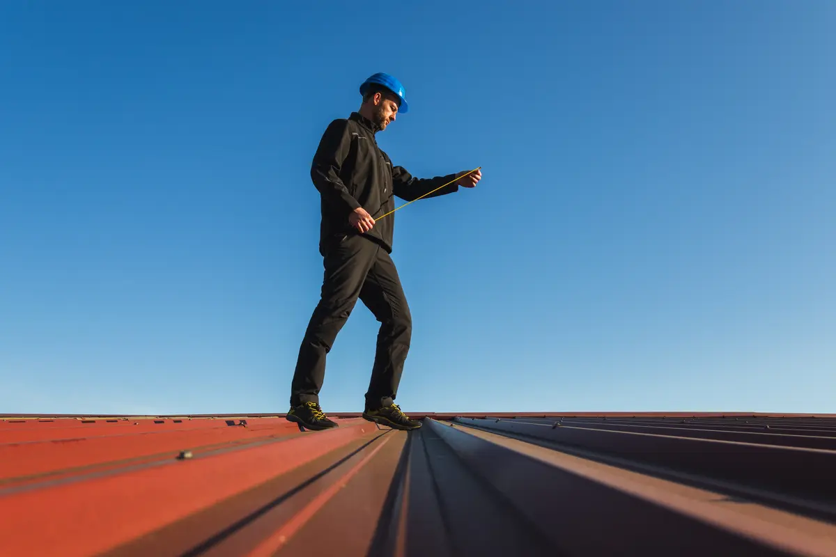 A man in dark clothing and a blue hard hat stands on a red metal roof, using a measuring tape to check the minimum slope for metal roof installation, with a clear blue sky in the background.