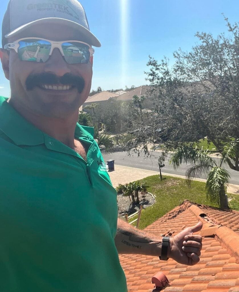 A man wearing sunglasses, a green polo shirt, and a cap stands on a red-tiled roof, smiling and giving a thumbs up. Trees, houses, and a bright sunny sky are visible in the background.