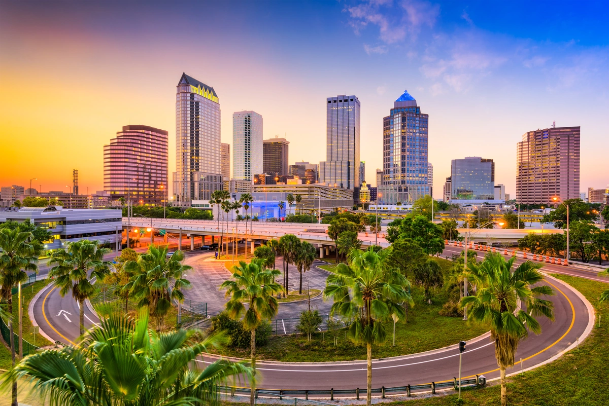 Downtown Tampa, Florida skyline at sunset features tall modern buildings, iconic Tampa landmarks, palm trees, and a curving road in the foreground beneath a vibrant, colorful sky.