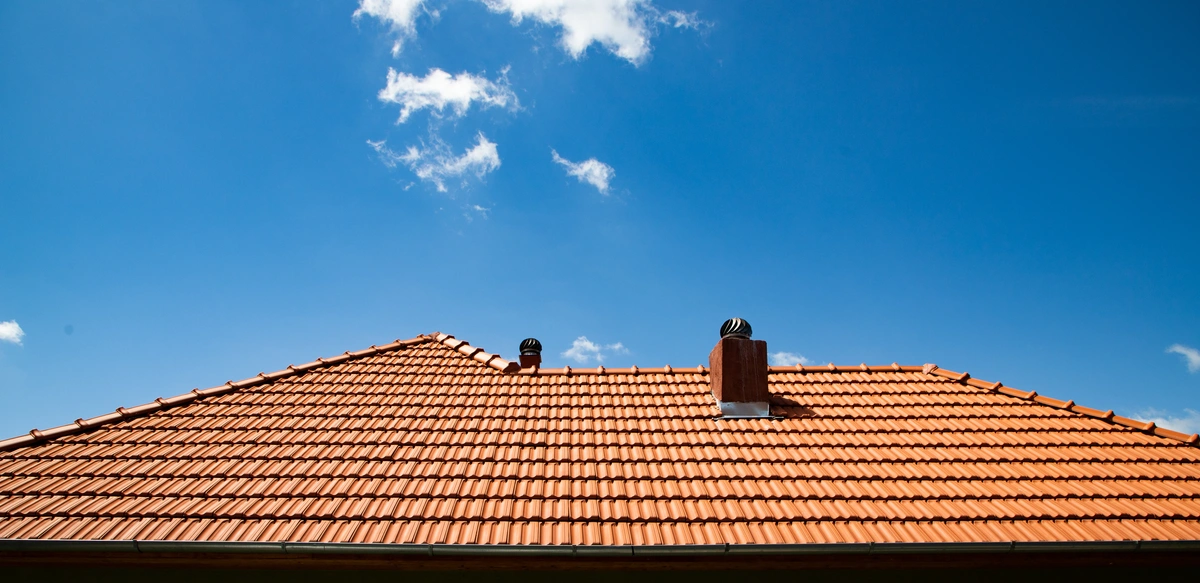 A red tiled roof with two small vents and a chimney sits under a clear blue sky with a few scattered white clouds, raising the question: how long does a tile roof last beneath such beautiful conditions?.