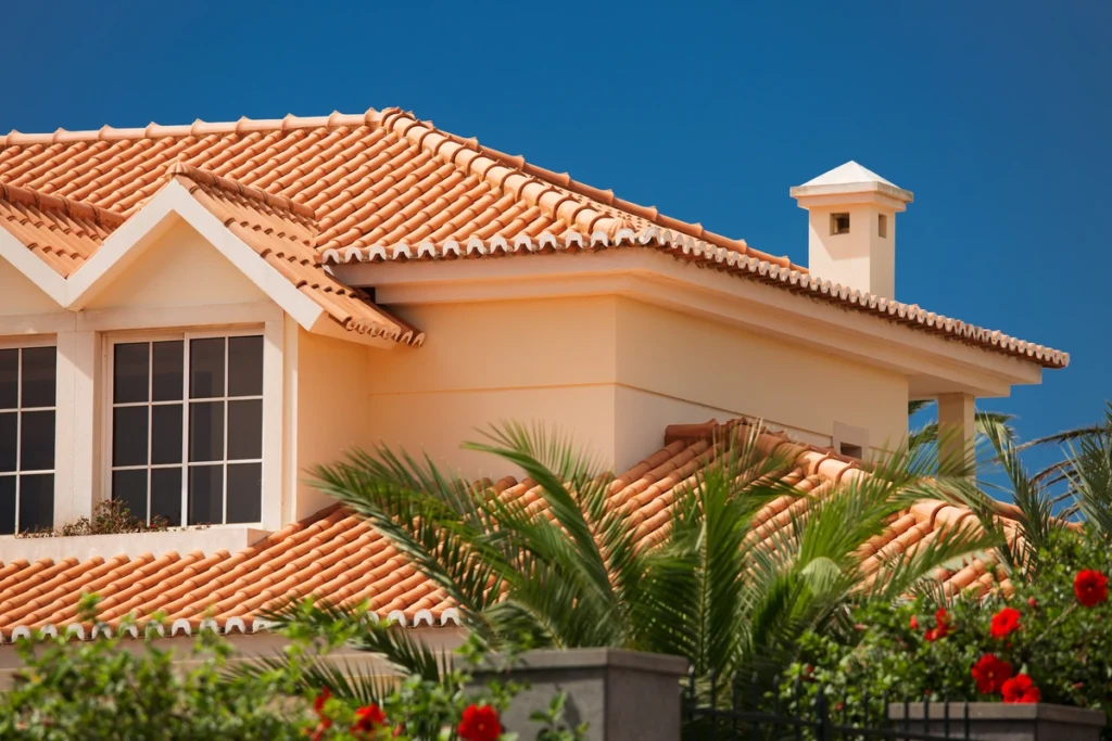 A modern house with orange tiled roof, large windows, and cream-colored walls—expertly crafted by roofing Polk County pros—surrounded by green palm leaves and red flowers under a clear blue sky.