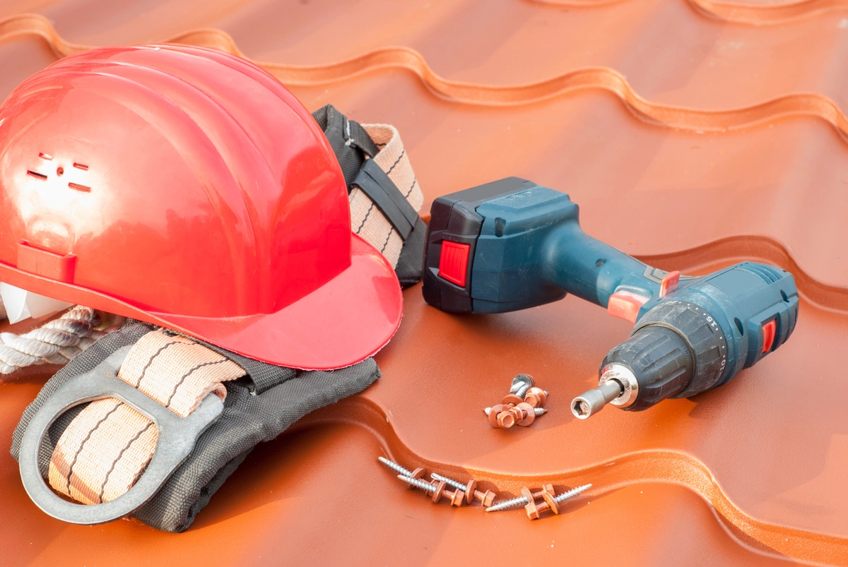 A red hard hat, a safety harness, an electric drill, and several screws—essential roofing tools—are placed on a brown metal roof, indicating preparation for construction or repair work.