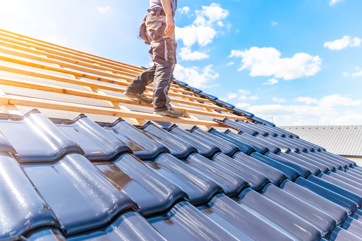A person demonstrates how to walk on tile roof while standing on a slanted roof under construction, with shiny blue tiles in the foreground and exposed wooden beams set against a bright, partly cloudy sky.