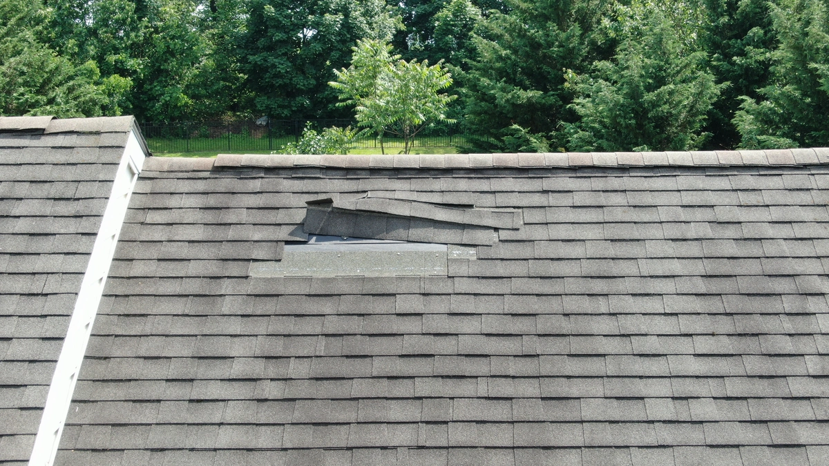 A house roof with several damaged and missing shingles, exposing the underlayment—clear signs of wind damage to roof. Trees and greenery are visible in the background.
