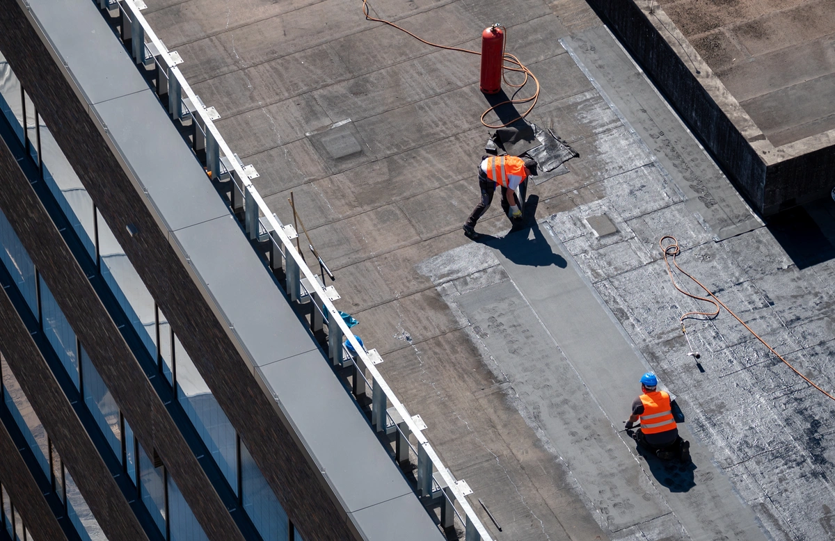 Aerial view of two construction workers in orange vests and hard hats working with flat roof materials on a rooftop; one is kneeling, and the other stands near tools and a red gas canister.