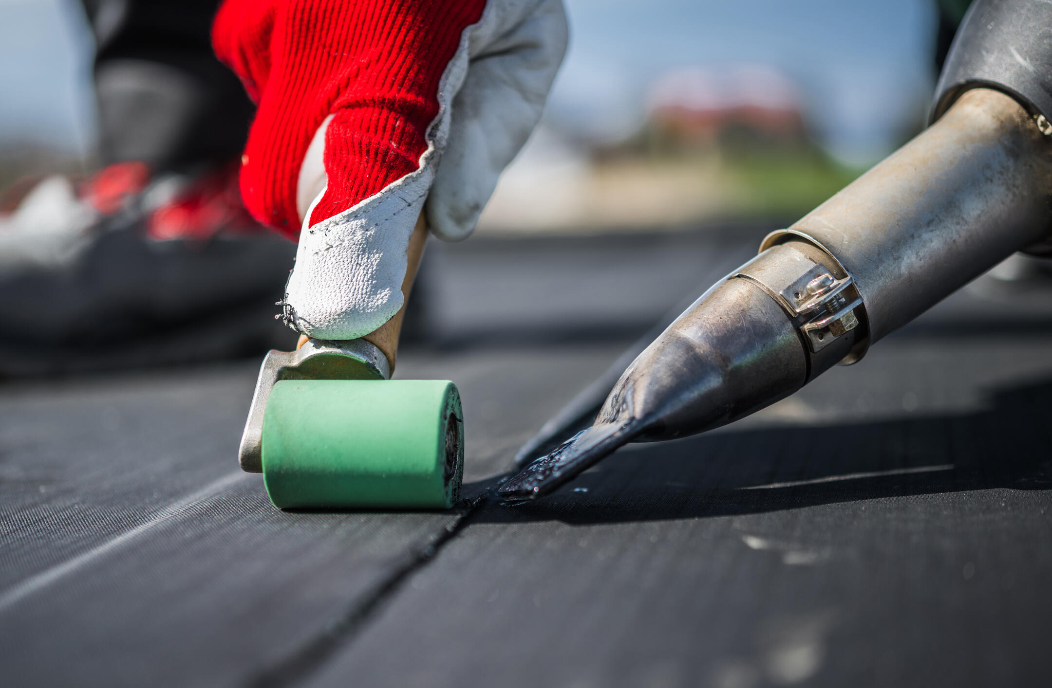 A close-up of a gloved hand holding a green tool while using a hot air gun to weld or seal a seam in a black synthetic roofing membrane, right next to rows of black shingles.