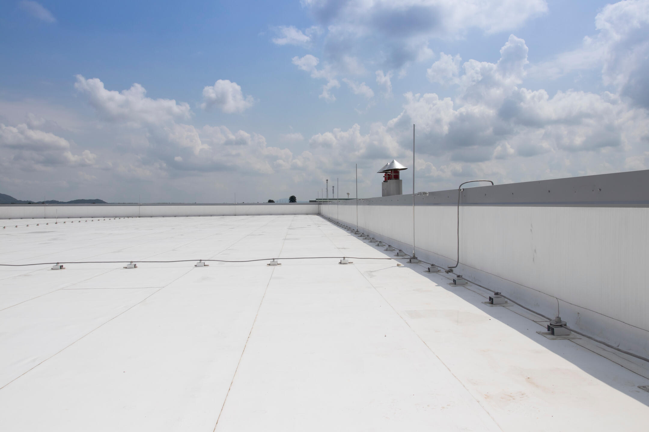 A large, flat, white rooftop with black shingles extends into the distance under a partly cloudy sky, bordered by a low wall on the right and with a small tower visible near the center background.