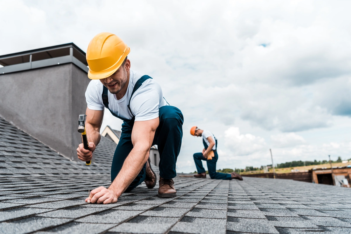 Two construction workers in yellow hard hats, equipped with roof safety equipment, are installing shingles on a sloped roof. One hammers in the foreground while the other works farther back. The sky is cloudy and buildings can be seen in the background.