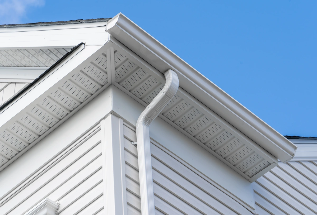 Close-up view of a white house’s exterior showcases the roofline, soffit, and downspout attached to the gutter, with drip edge flashing visible against a clear blue sky.