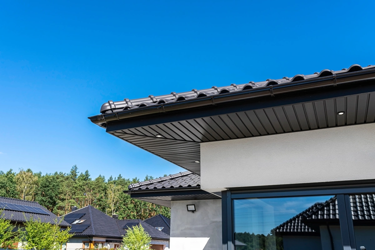 Modern house with a dark tiled roof and sleek roof eaves, black gutters, and white exterior walls, set against a clear blue sky and surrounded by trees and neighboring houses.