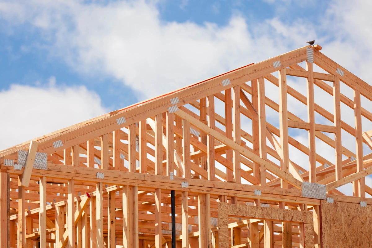 Wooden framework of a house under construction, highlighting exposed roof trusses set against a blue sky with scattered clouds.