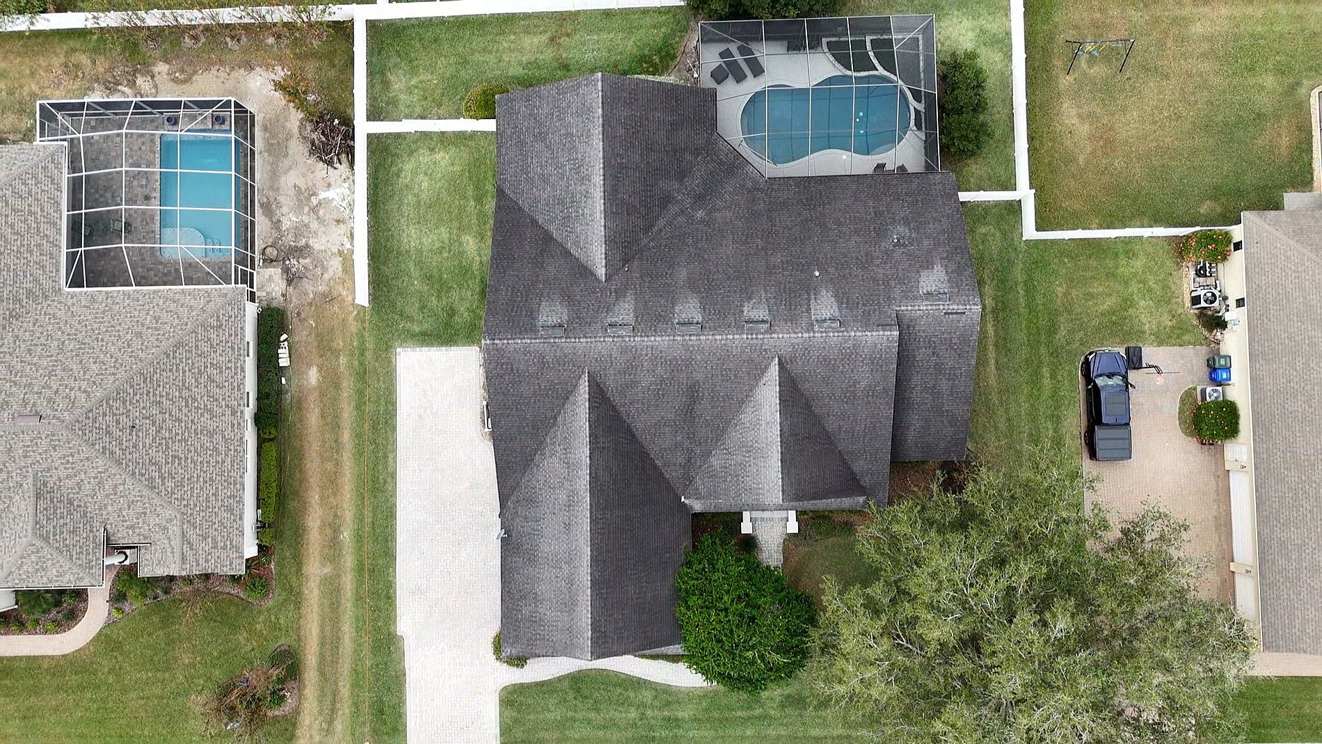 Aerial view of a house with a dark roof, driveway, and backyard pool enclosed in a screened structure. Neighboring houses and lawns, another pool, a driveway with parked vehicles, and a swing set are also visible.