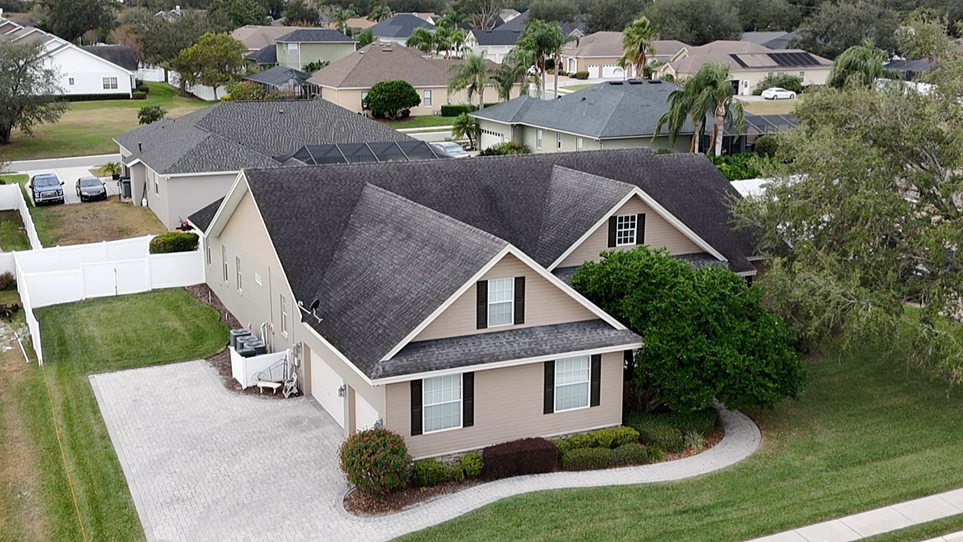Aerial view of a beige house with a dark roof, white trim, and landscaped yard in a suburban neighborhood, surrounded by other homes and greenery.