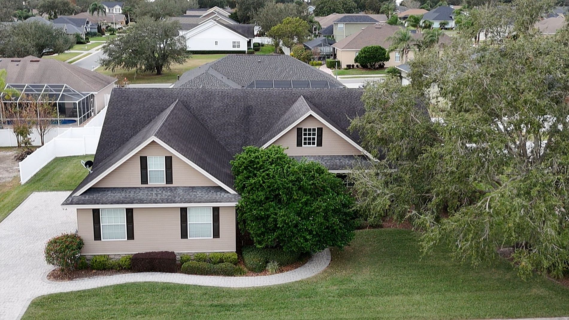 Aerial view of a suburban house with a dark roof, beige siding, white trim, and a curved driveway, surrounded by a well-maintained lawn, shrubs, and neighboring houses.