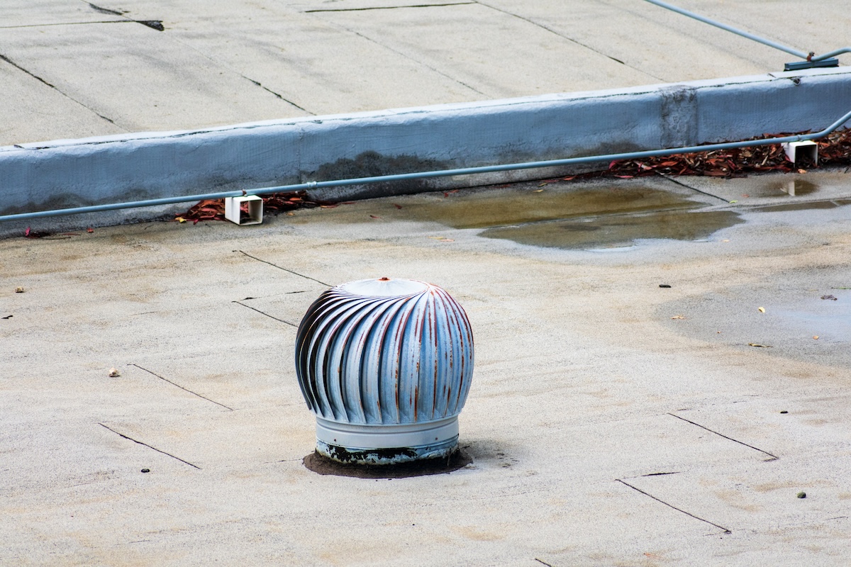 A silver metal turbine roof vent sits on a flat, beige rooftop. Scattered leaves, some puddles—signs often warranting flat roof leak repair—are present, along with a low concrete barrier in the background.
