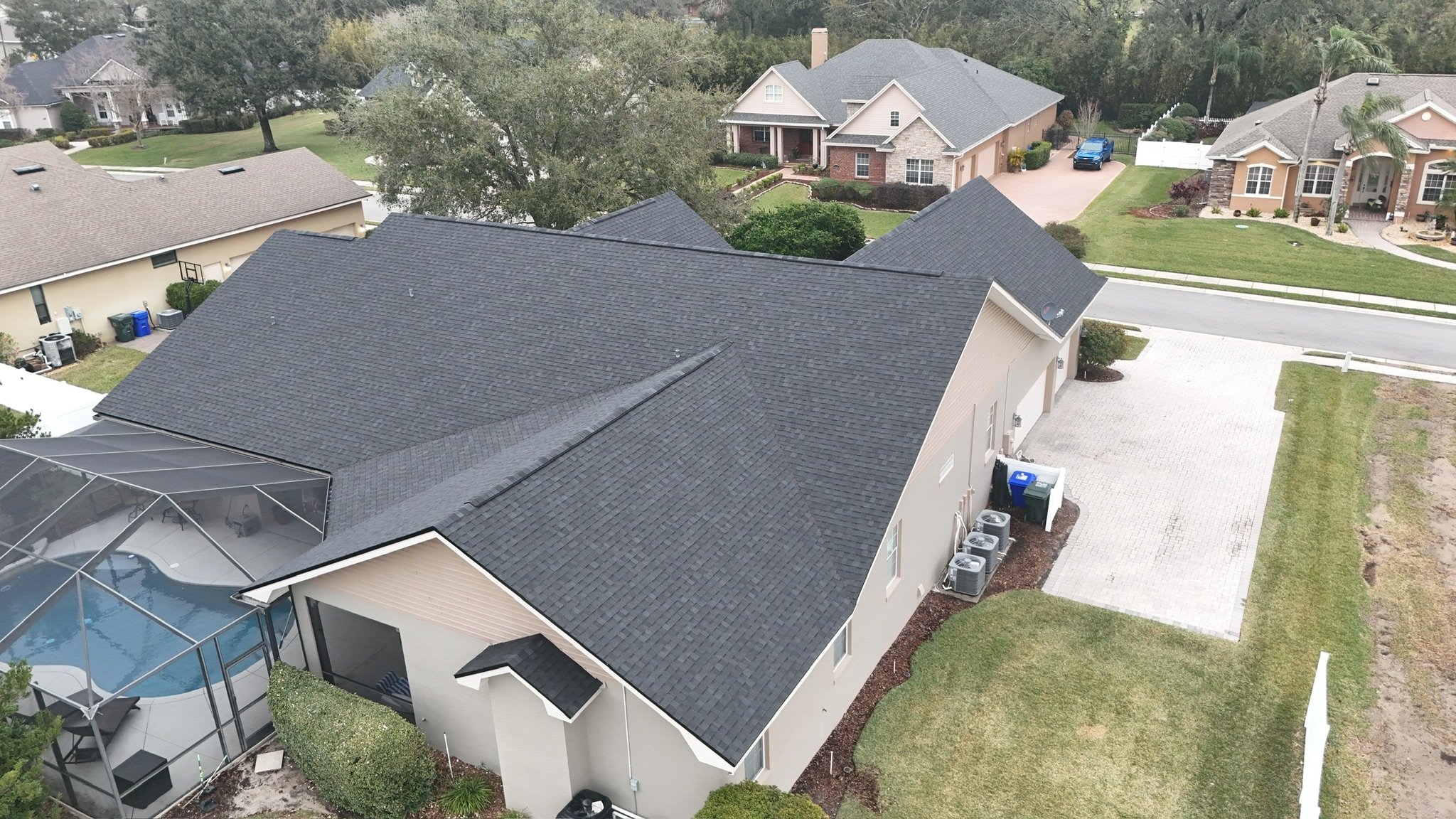 Aerial view of a suburban house with a dark gray shingle roof, attached screened pool area, and well-kept yard, surrounded by other homes and green lawns in a residential neighborhood.