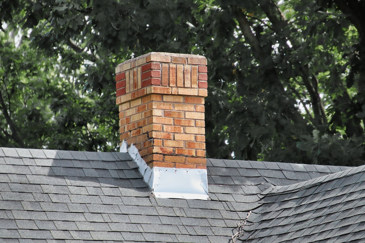 A brick chimney extends above a shingled roof, surrounded by expertly installed metal roof flashing repair. In the background, leafy green trees are visible.