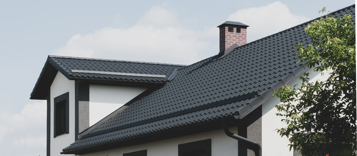 A modern house with a dark, tiled sloped roof, showcasing expert tile roof maintenance, features white walls accented with dark trim, a red-brick chimney, and a green tree beside the building under a partly cloudy sky.