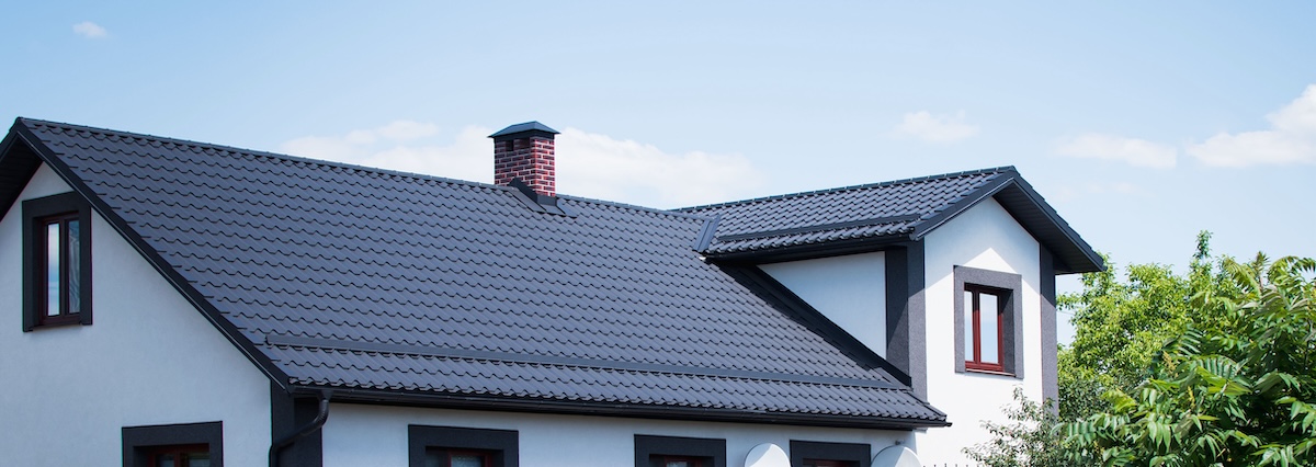 A modern house with white walls, dark-framed windows, and a sleek tile roof installation in dark gray metal. A red brick chimney sits atop the roof, surrounded by green trees under a clear blue sky.