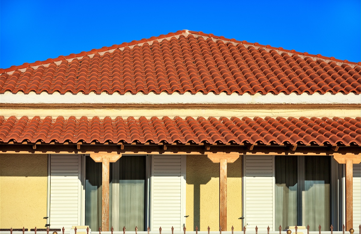A building with a red, tiled roof and cream-colored walls stands under a clear blue sky. Four windows feature white shutters and curtains. Wondering how long does a clay tile roof last? These roofs are known for their impressive durability.