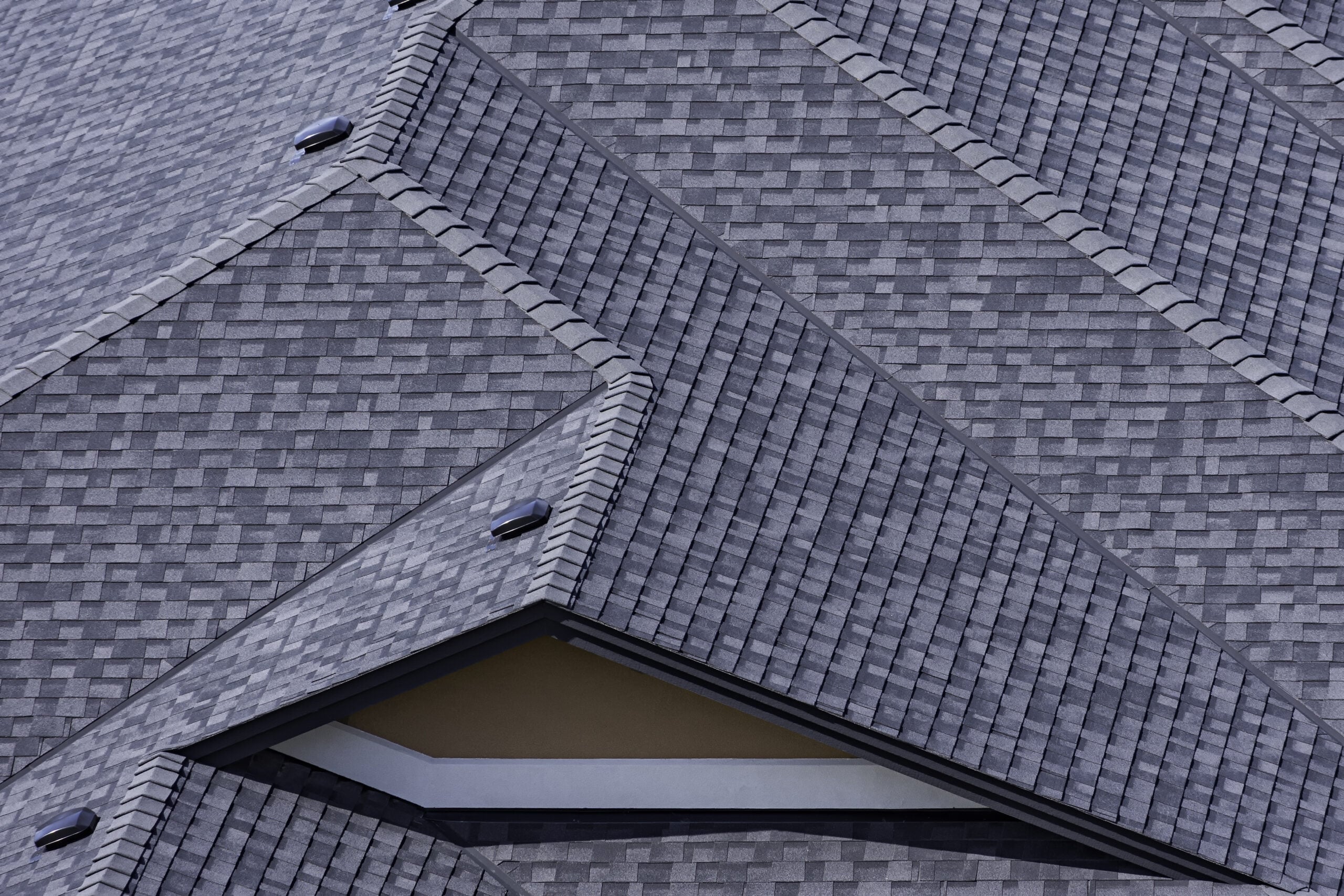 Gray asphalt shingles cover intersecting, multi-angled roof sections, creating a geometric pattern. Small vents and a discreet roof patch are visible on some ridges, while a section of beige wall peeks out below an overhang.