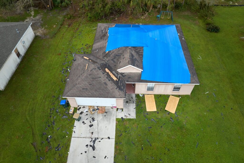Aerial view of damaged in hurricane Ian house roof covered with blue protective tarp against rain water leaking until replacement of asphalt shingles.
