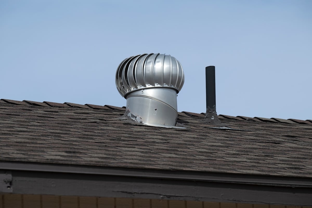 A close-up view of a rooftop showing a silver metal turbine vent and a black exhaust pipe—evidence of expert roof vent installation—protruding from brown asphalt shingles under a clear blue sky.