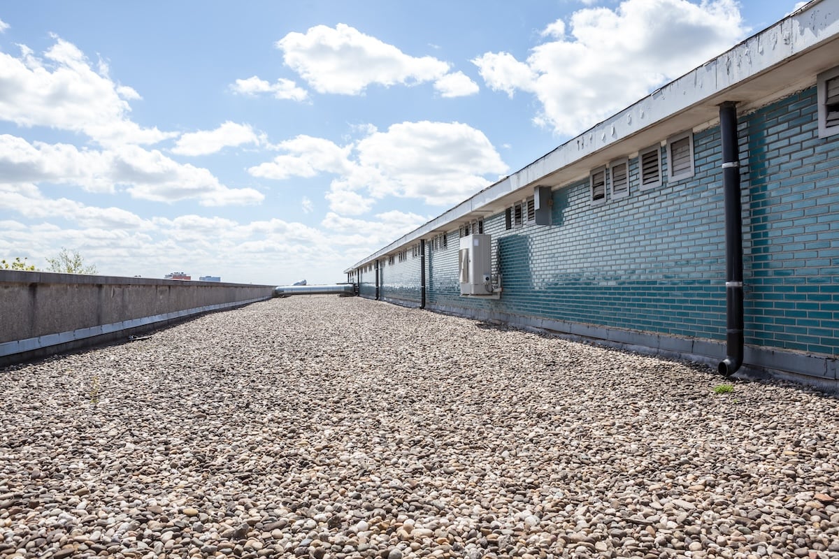 Flat tar and gravel roof covered with small stones, bordered by a low concrete wall on the left and a long building with blue-green tiles and pipes on the right, under a partly cloudy sky.