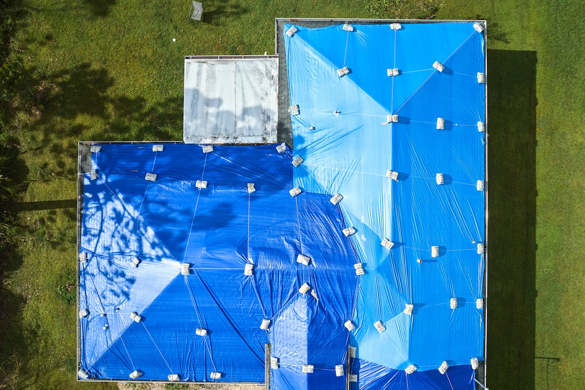 Aerial view of a house roof covered with an emergency roof tarp, secured by numerous concrete blocks, surrounded by green grass and some trees casting shadows.
