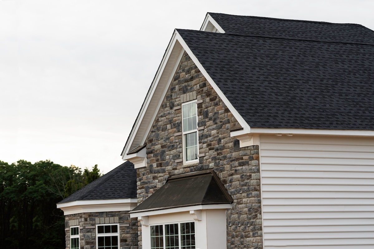 A close-up view of a modern house exterior with gray stonework, white siding, black roof shingles, and large windows. Trees are visible in the background under a cloudy sky.