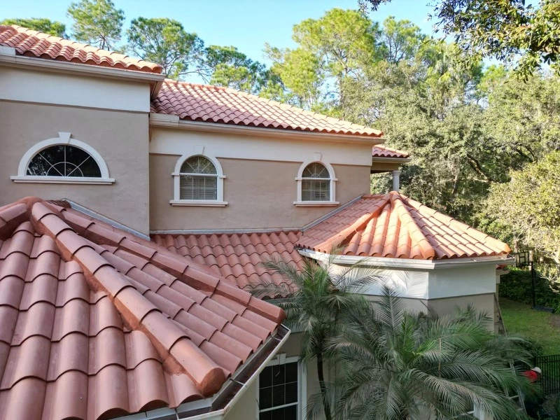 A tan stucco house with red clay tile roofing by roofing Port Charlotte, arched windows with white trim, and palm trees in front, surrounded by tall green trees under a clear blue sky.