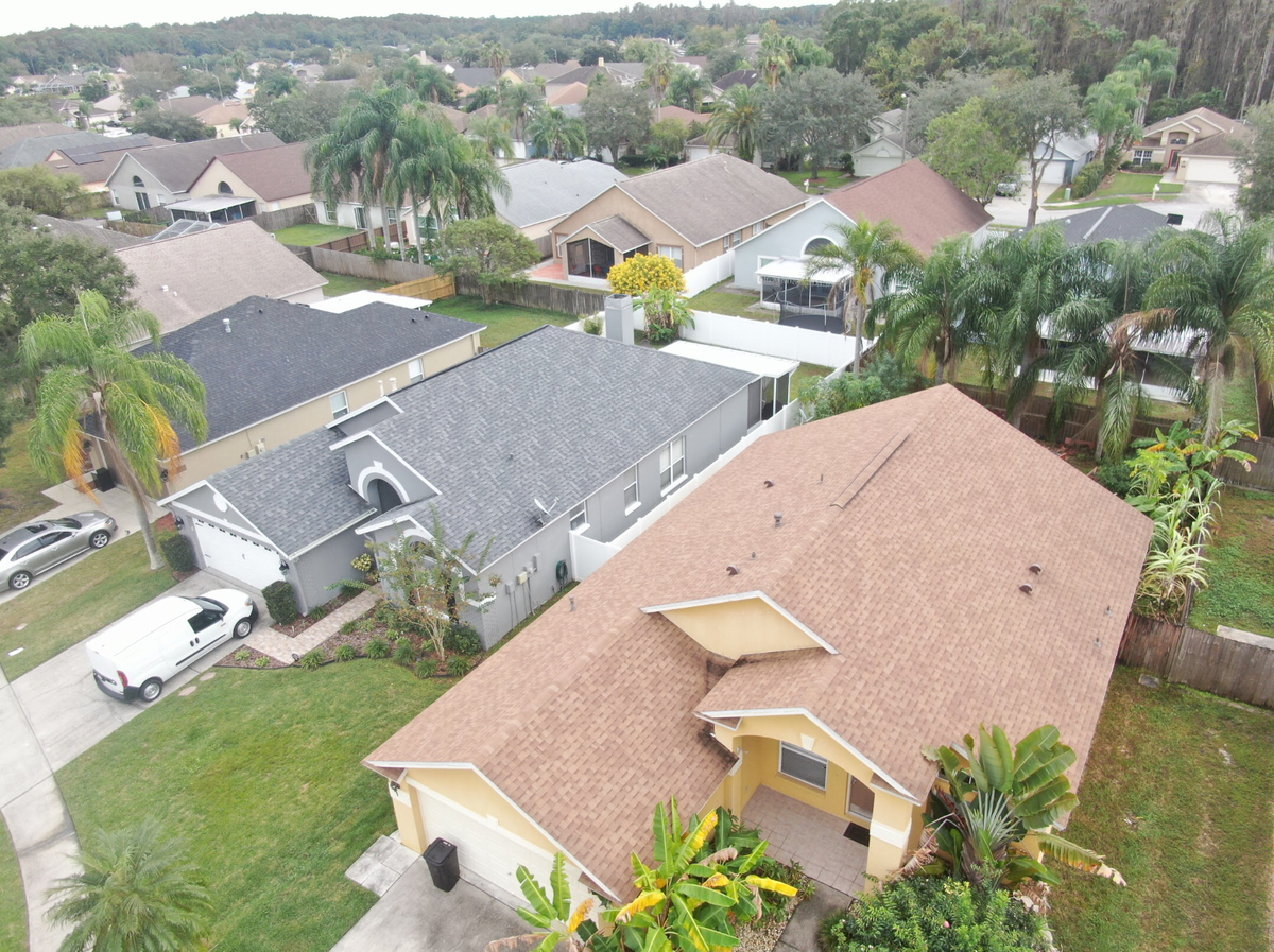 Aerial view of a suburban neighborhood in Valrico with single-family homes, green lawns, palm trees, and driveways. The roofs display various shades of brown and gray—showcasing the quality of roofing Valrico residents trust. A white van is parked in one driveway.